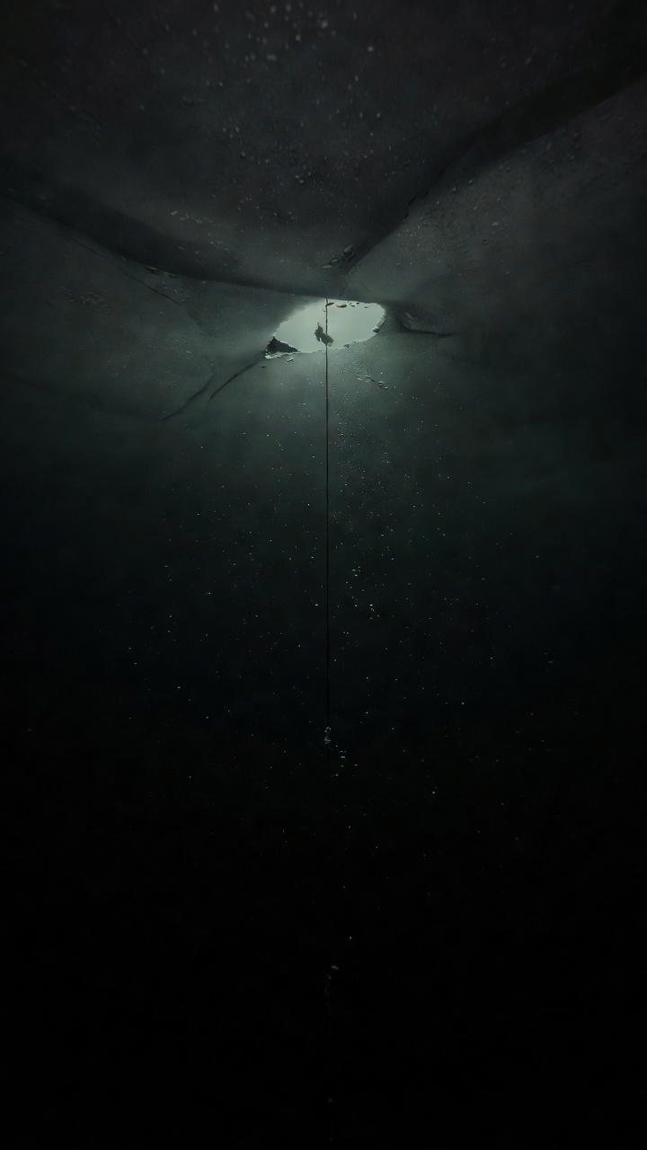Looking up through black water at a small ragged hole in lake ice, a single dive line dropping from it, a tiny diver silhouette near the surface, the depths below fading to black.