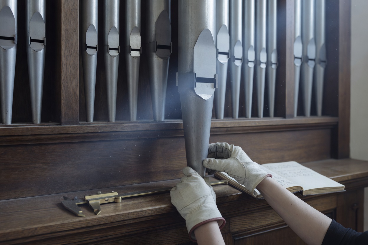 Silver organ facade pipes from below, gloved hands lifting one pipe from its rack, a caliper and ledger on the gallery rail in cold light.